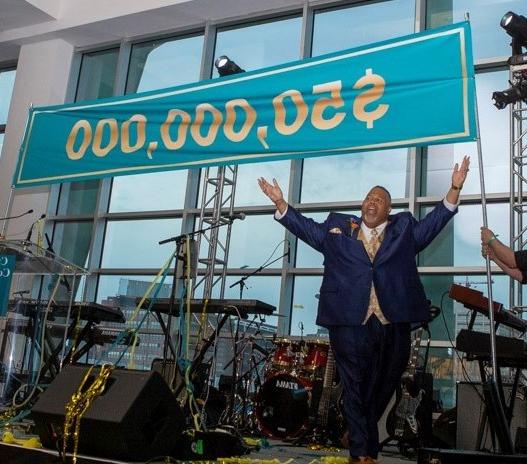 Dr. Michael Baston, arms raised, on stage in front of banner announcing $50 million goal achieved